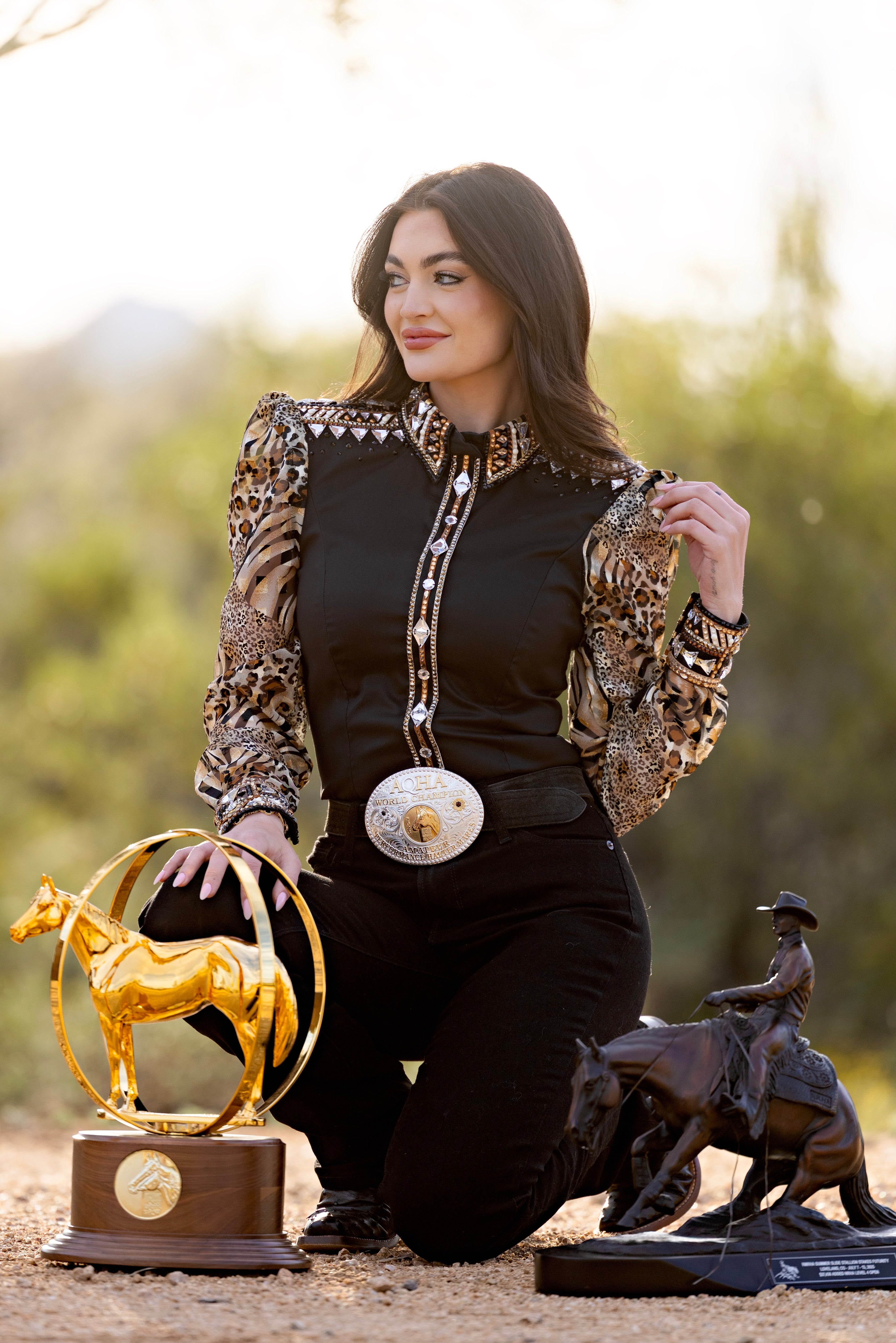 Woman in a stylish outfit with decorative elements, kneeling next to a gold and bronze sculpture outdoors.